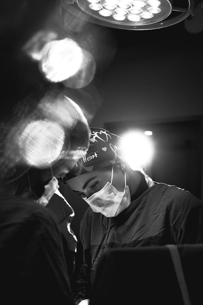 A focused surgeon in a surgical mask and cap performing an operation in a black and white operating room setting.